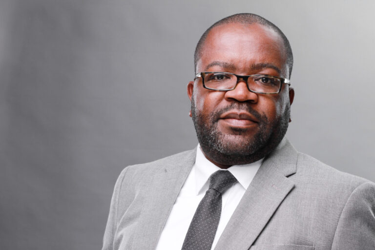 Headshot of Tutaleni Asino wearing a gray suit jacket and a dark tie. He is also wearing glasses and sporting a beard.