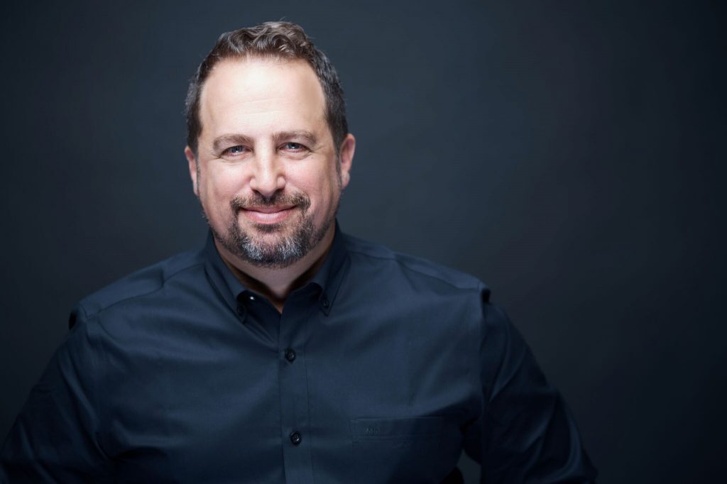 Headshot of Gad Barnea wearing a black button-down shirt against a dark background.