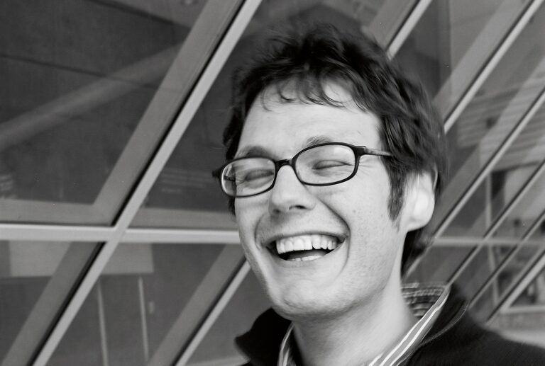 A headshot of Ilya Kaminsky, eyes closed, black glasses, laughing.