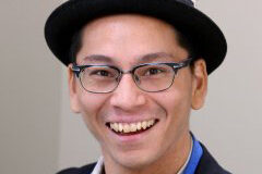 A headshot of Kian Mintz-Woo in a black hat and blue-rimmed glasses.