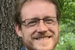 Headshot of Scott Duxbury wearing glasses, sporting a goatee and posing in front of tree.