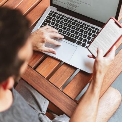 A person working on a laptop while looking at their cell phone.