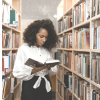 A Black woman in the library stacks, reading a book.